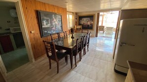 A dining area with a glass table and wooden chairs, a television mounted on the wall, and a refrigerator in the corner. - Vintage Rooftop (Heliopolis)