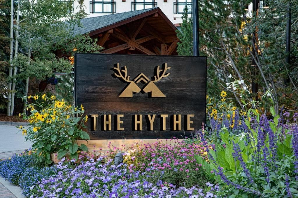 A wooden sign with a logo and the text 'THE HYTHE' in front of a building with a sloped roof and wooden beams.