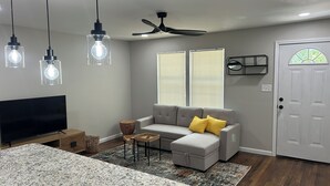 A modern living room with a grey sofa, ottoman, and a coffee table. There is a TV, a ceiling fan, and a door with a window. - Cozy 1-bedroom in charming Durham (Durham)