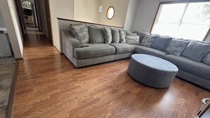 A modern living room with a grey sectional sofa, a grey ottoman, and a wooden floor. - Harmony Haven (Saint Robert)