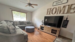 A living room with a grey sofa, ottoman, wooden floor, ceiling fan, TV, and a 'HOME' sign on the wall. - Harmony Haven (Saint Robert)
