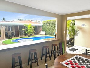 A room with a view of a swimming pool, a table with a checkered table runner, and a bar area with stools. - Private residential home for 8 with private pool; 5 min walk to Jacó Beach. (Jaco)