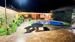 A swimming pool with a patio area, surrounded by greenery and a building with a covered seating area.