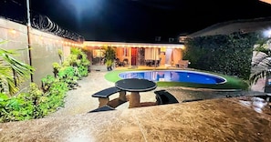 A swimming pool with a patio area, surrounded by greenery and a building with a covered seating area.