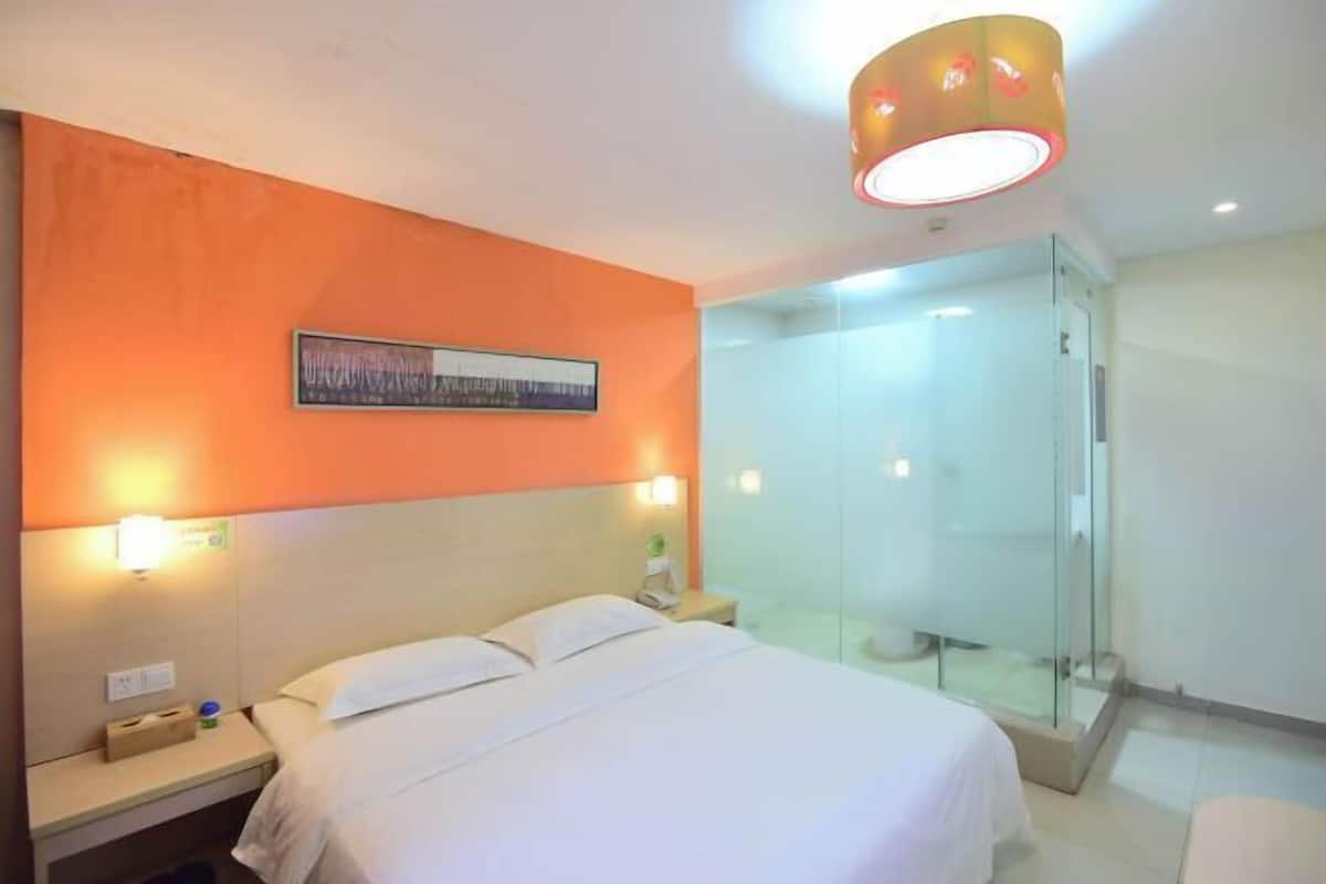 A hotel room with a bed, a nightstand, a wall-mounted light, a glass shower enclosure, and a framed picture on the wall.