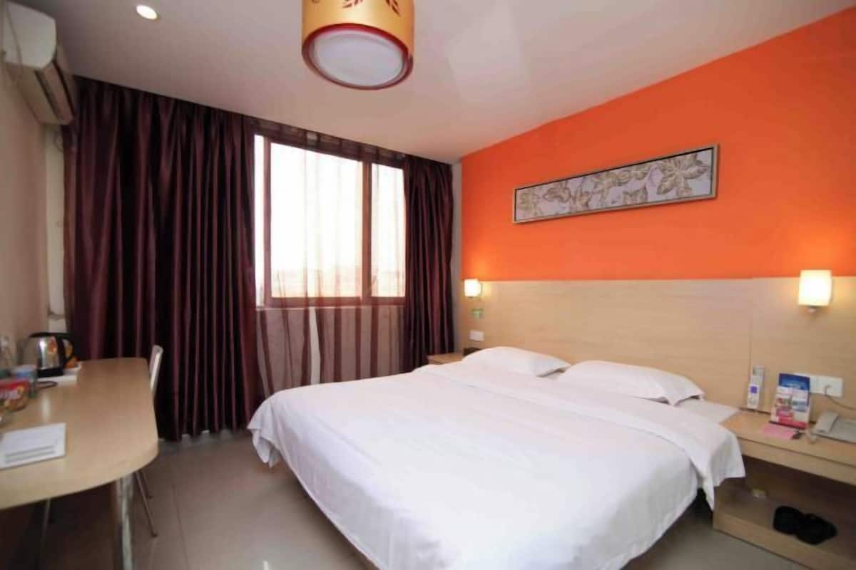 A hotel room with a bed, a desk, a chair, a window with curtains, a wall-mounted artwork, and a nightstand with a lamp.