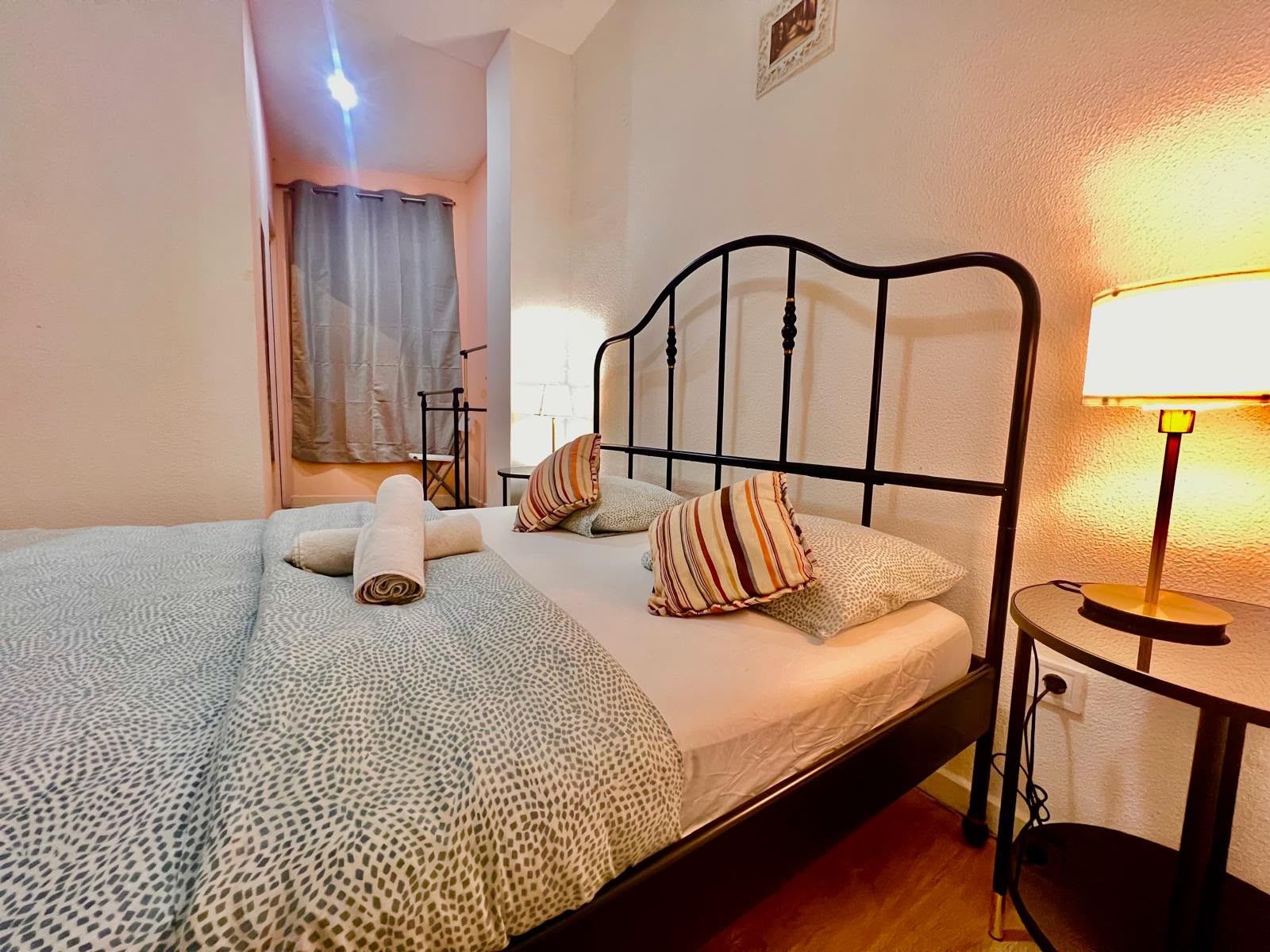 Una cama bien hecha con una colcha estampada, dos almohadas y una toalla enrollada. Hay una lámpara de noche sobre una mesita junto a la cama.