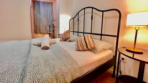 Ein ordentlich bezogenes Bett mit einem gemusterten Überwurf, Kissen und einem zusammengerollten Handtuch. Ein Nachttisch mit Lampe und Steckdose ist vorhanden.