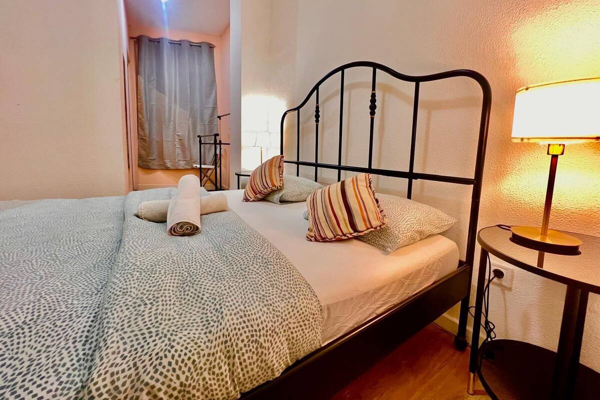 A neatly made bed with a patterned bedspread, pillows, and a rolled towel. There is a bedside table with a lamp and a power outlet.