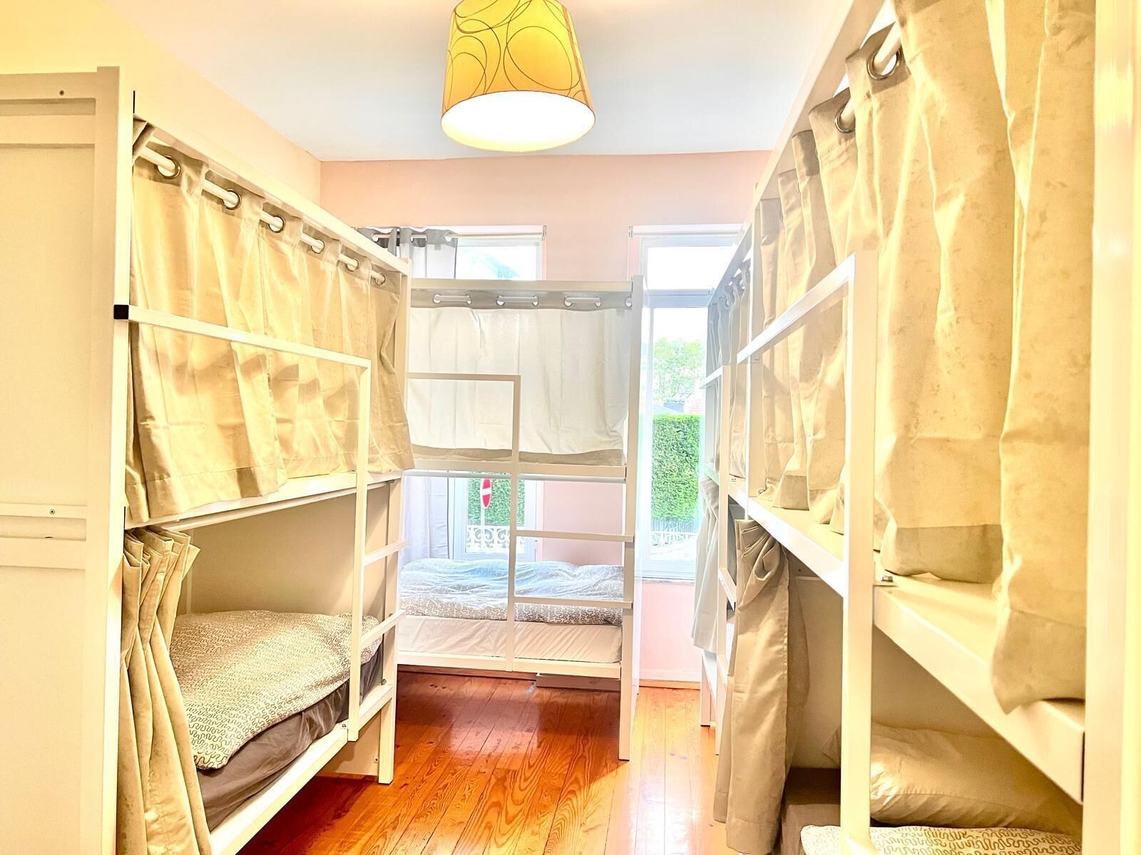 A dormitory room with bunk beds, a wooden floor, and a window with curtains.