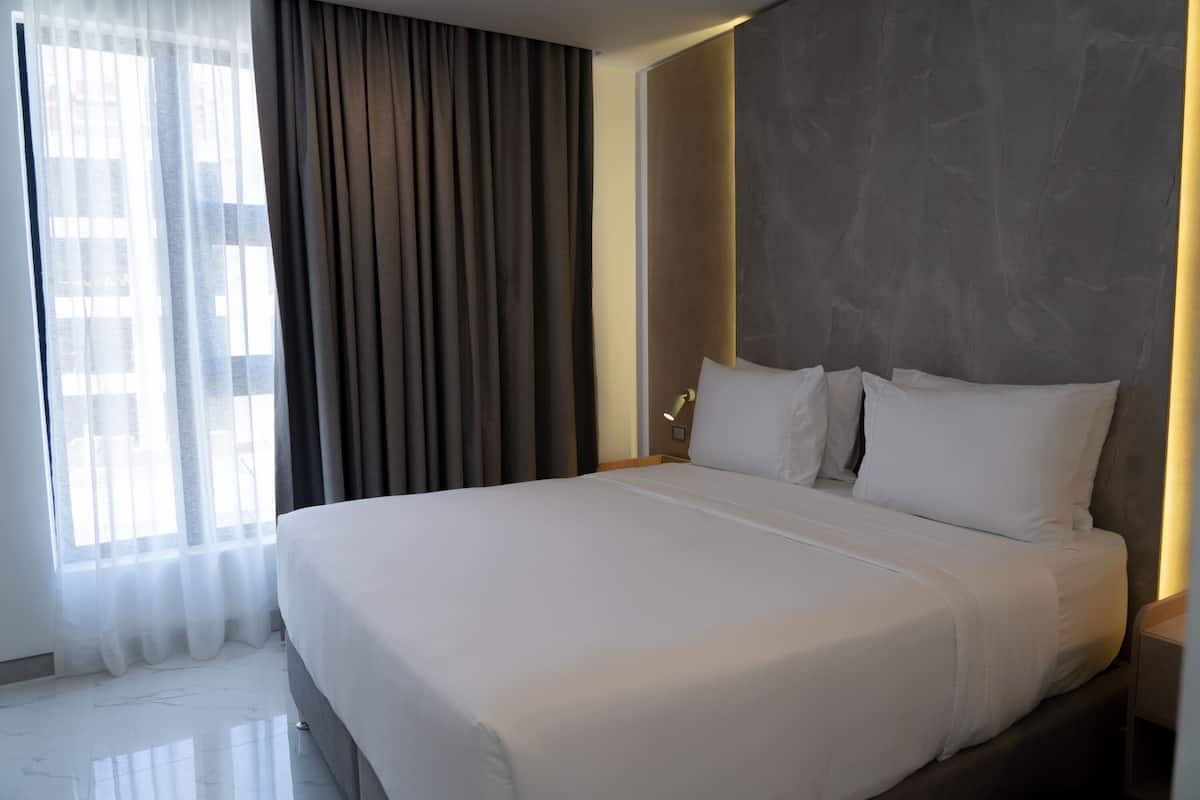A modern hotel room with a large bed, a nightstand, a wall-mounted lamp, and a window with sheer curtains.