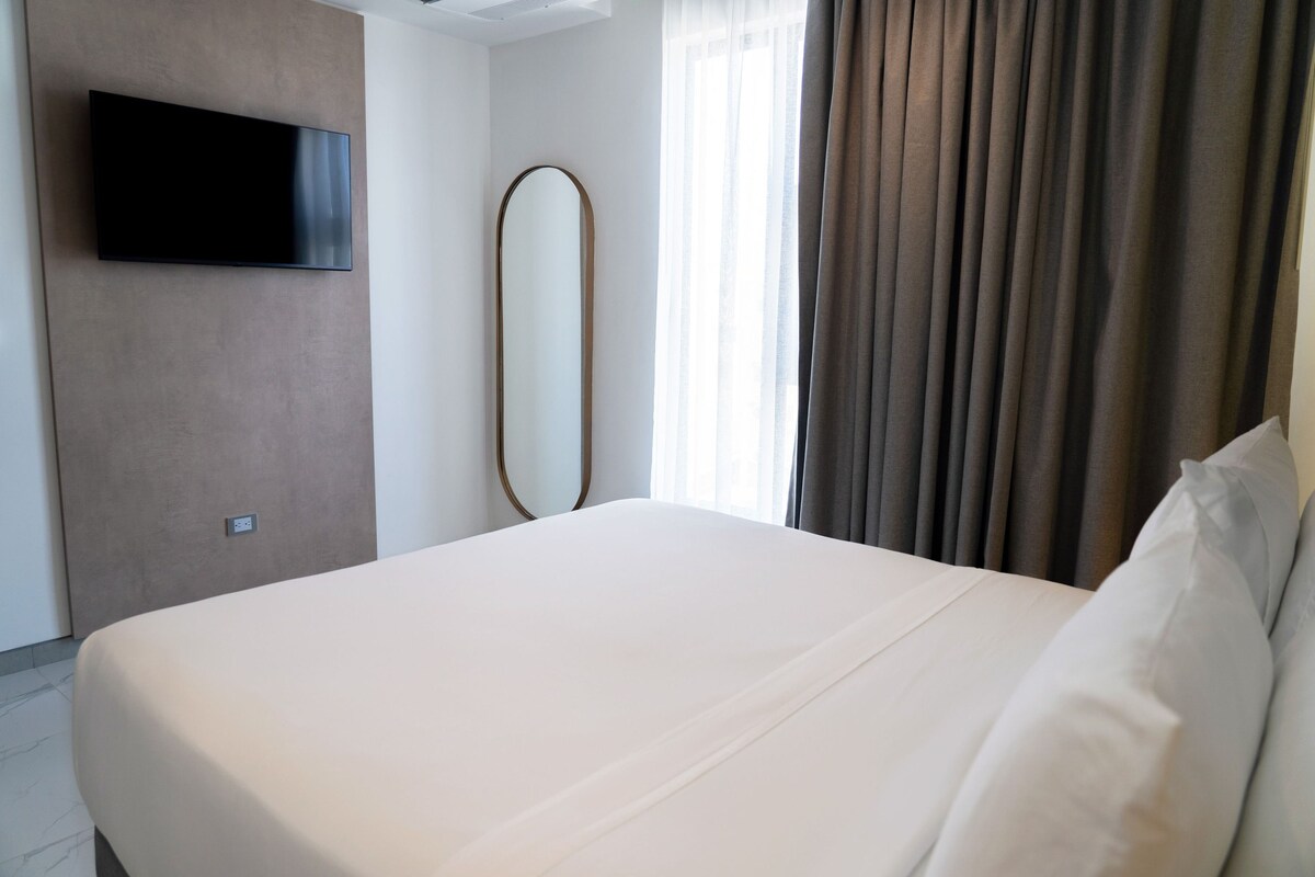 A hotel room with a bed, a flat-screen TV mounted on the wall, a large mirror, and a window with curtains.