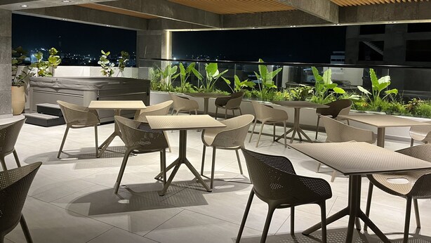 Un patio al aire libre con varias opciones de asientos, incluyendo mesas y sillas, rodeado de plantas en macetas y vistas de la ciudad de noche.