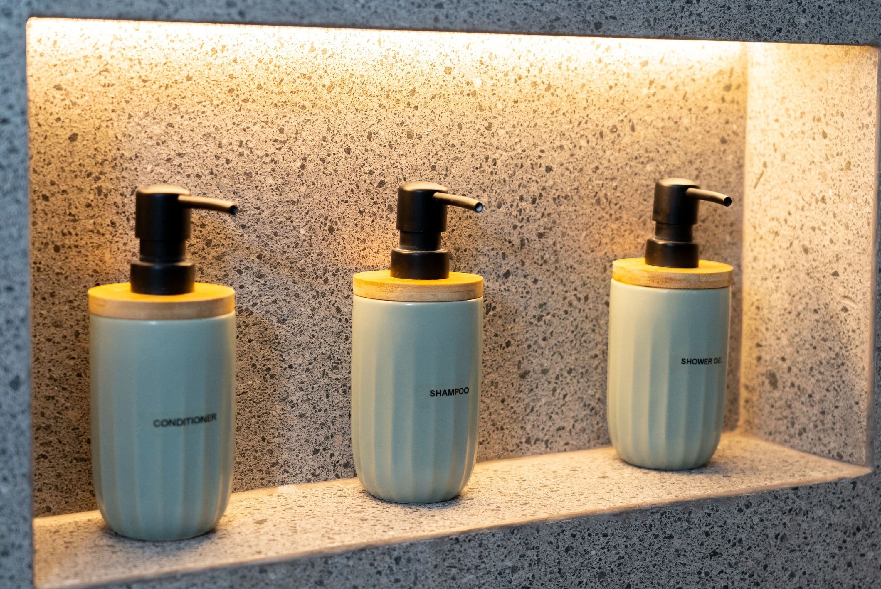 Three bottles of shampoo and conditioner on a shelf against a speckled wall.