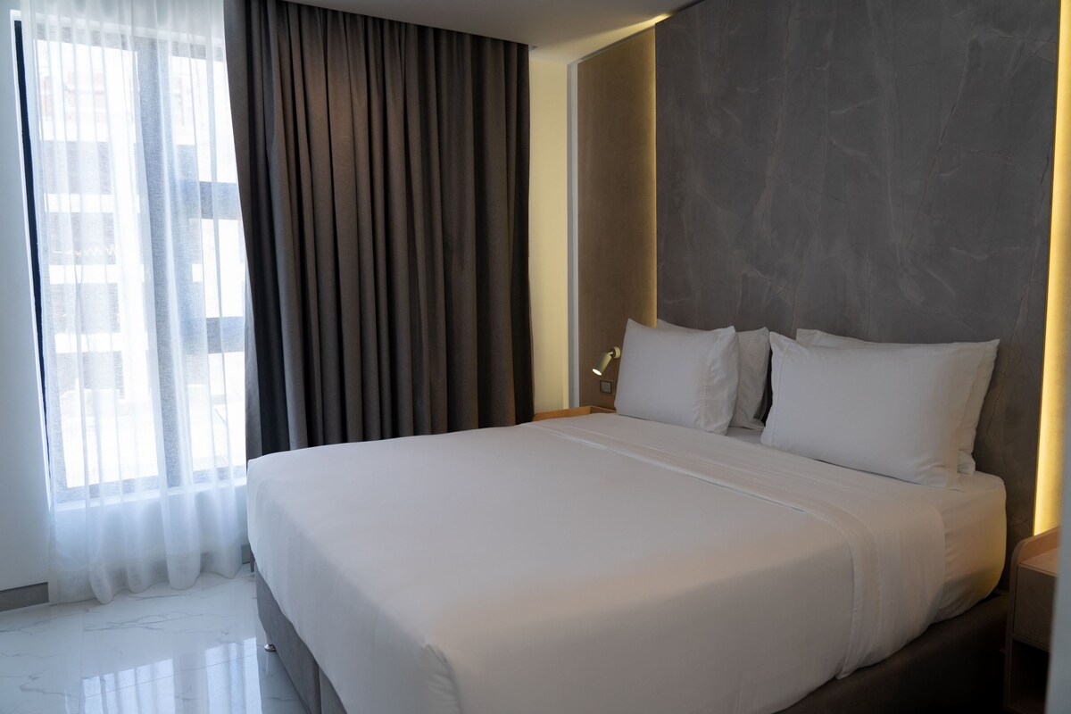 A modern hotel room with a large bed, a nightstand, a wall-mounted lamp, and a window with sheer curtains.