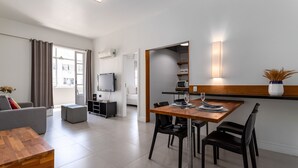Un salon moderne avec un canapé, une table basse en bois et un téléviseur. On y trouve également un coin repas avec une table et des chaises, ainsi qu’une cuisine équipée d’un micro-ondes et d’étagères.