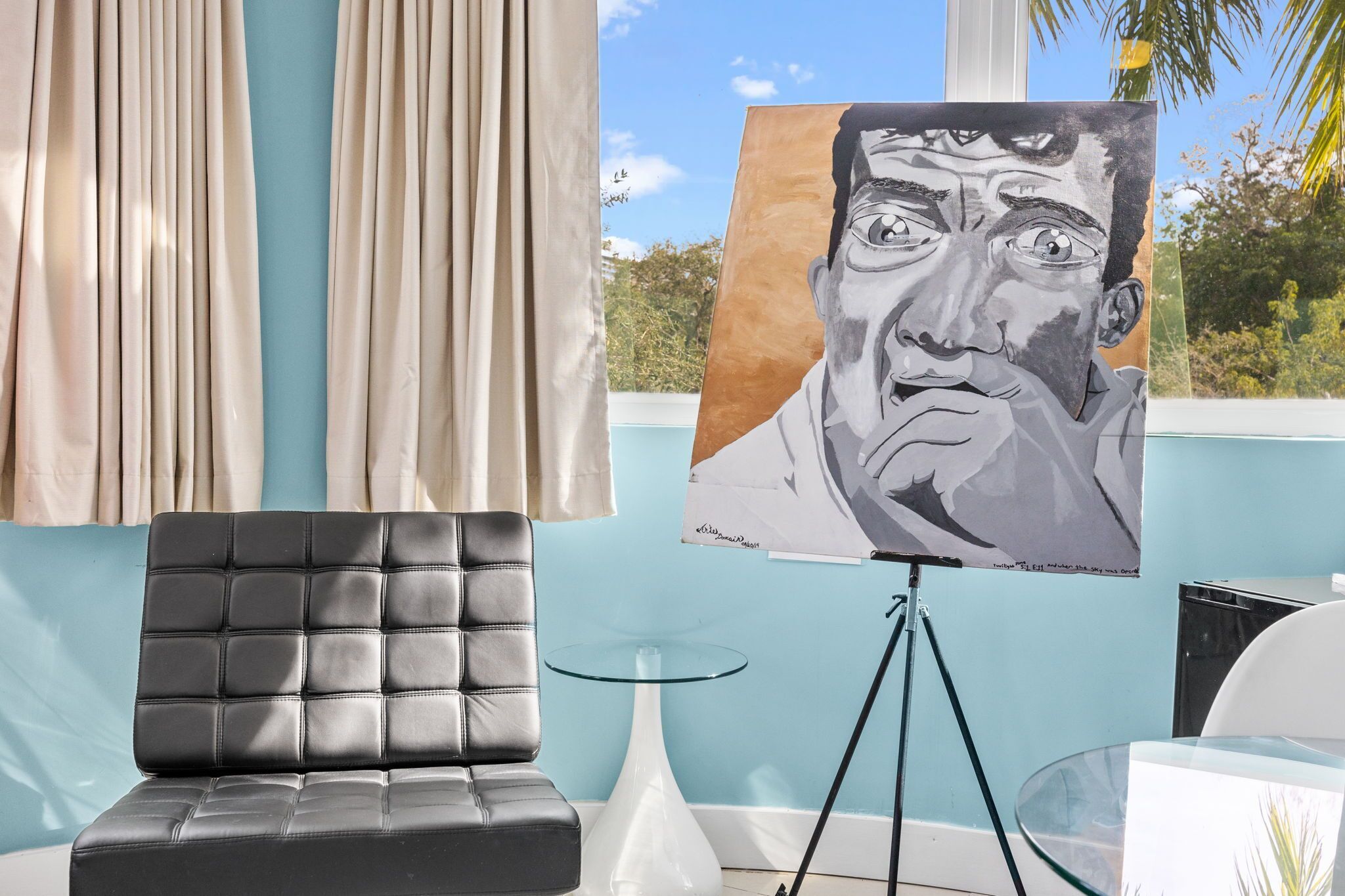 A room with a black leather sofa, a glass table, and a painting on a tripod stand.