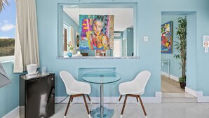 Ein modernes Zimmer mit einem Glastisch, zwei weißen Stühlen, einer schwarzen Minibar und einem großen Spiegel, der ein farbenfrohes Gemälde reflektiert.