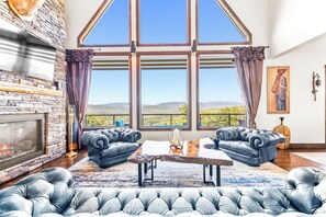 A spacious living room with a fireplace, two leather sofas, a coffee table, and a large window offering a scenic view. - Luxurious Spacious 6-bedroom House. Best Views, Hot tub. Waterville Estates!  (Campton)