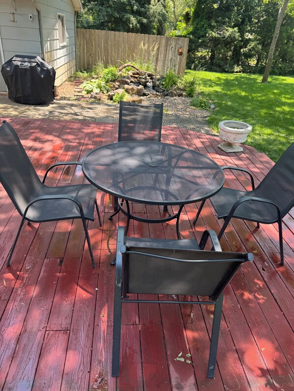 A patio with a round metal table and four chairs, surrounded by a wooden deck and a wooden fence.