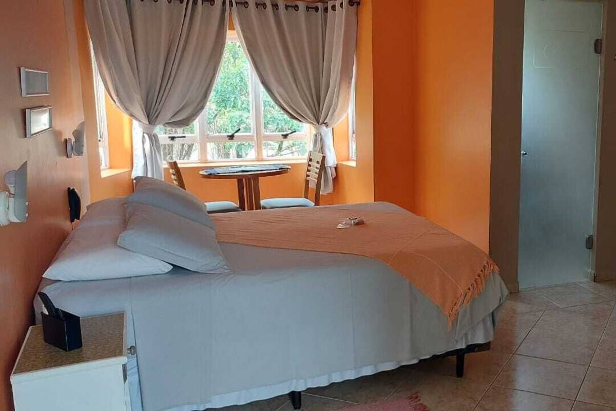 A bedroom with a bed, a small table, a chair, a window with curtains, and a bathroom with a sink and mirror.
