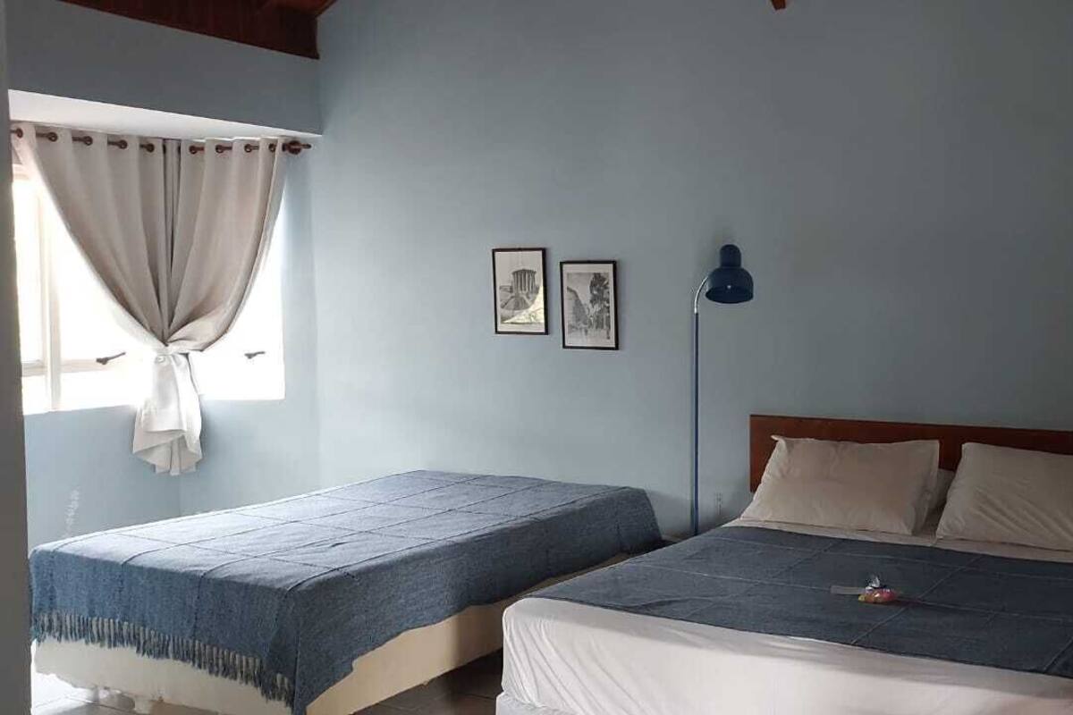 A room with two beds, a window with curtains, a lamp, and two framed pictures on the wall.