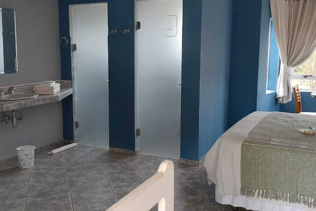 A bedroom with a bed, a bedside table, a window with curtains, a bathroom with a sink and mirror, and a door.
