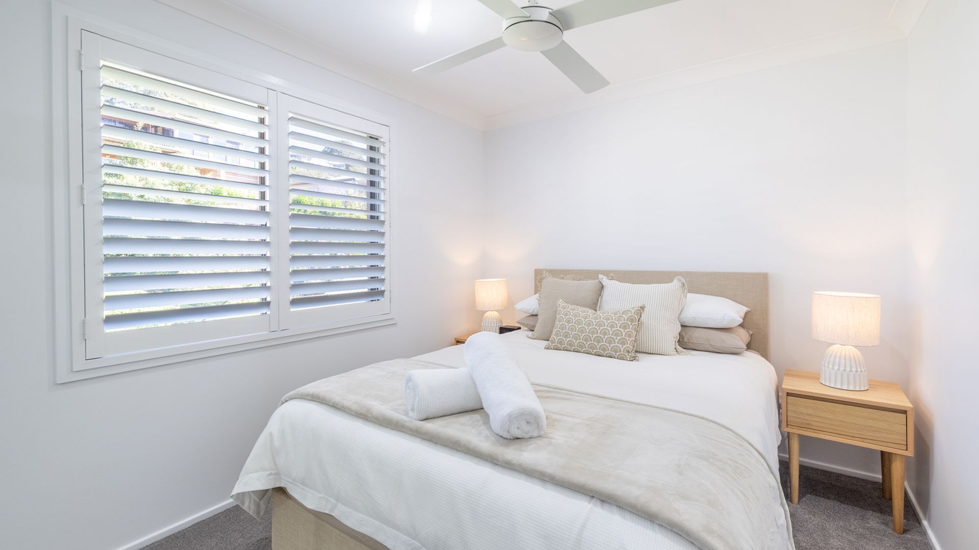 Un dormitorio con cama, dos mesitas de noche, ventilador de techo y ventana con persianas.