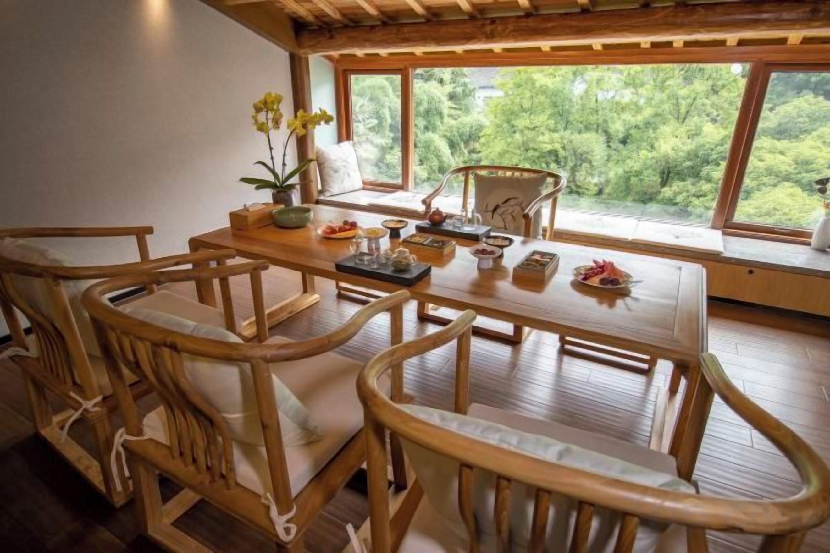 Un comedor con mesa y sillas de madera, un florero con flores y vistas a zonas verdes a través de amplios ventanales.