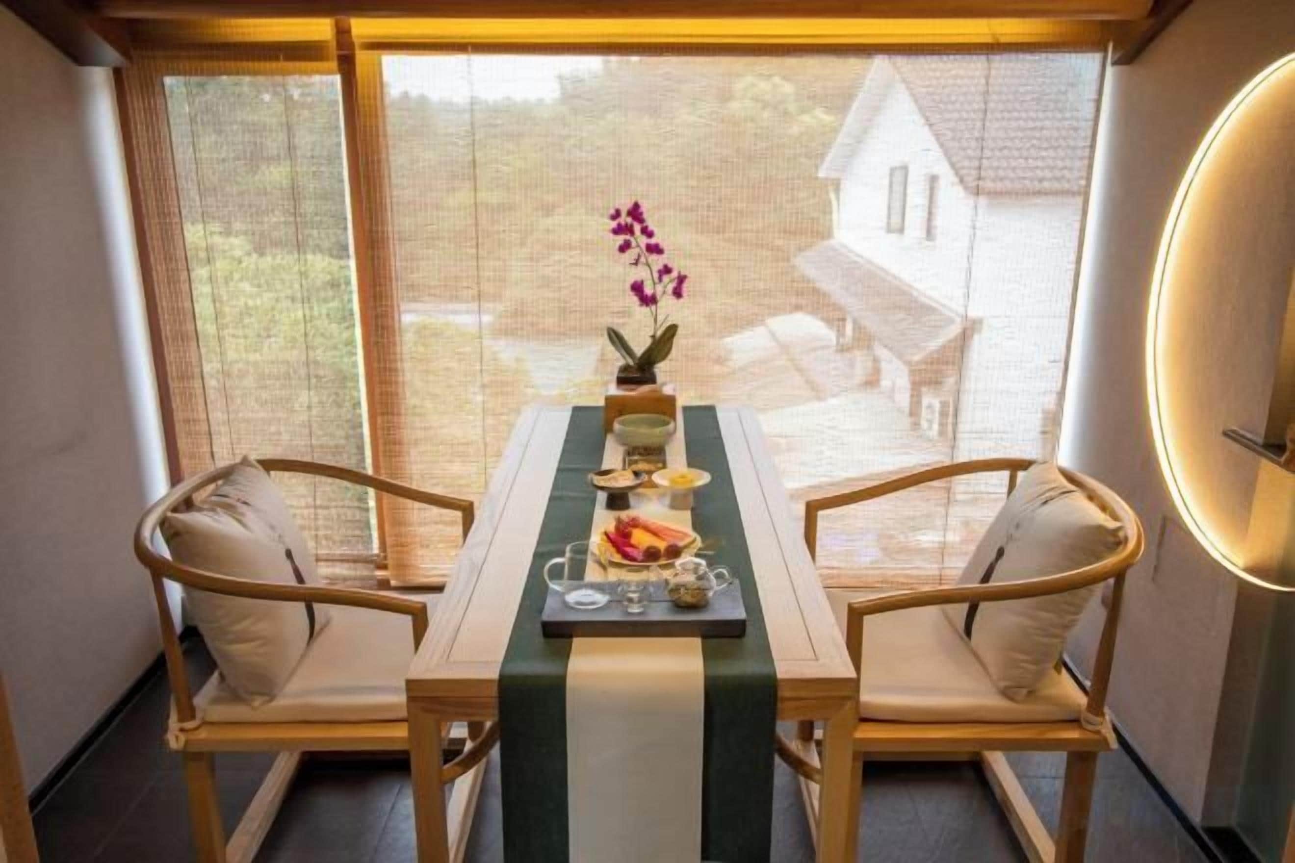A dining area with a table set for two, a vase with flowers, and a view of the outside.