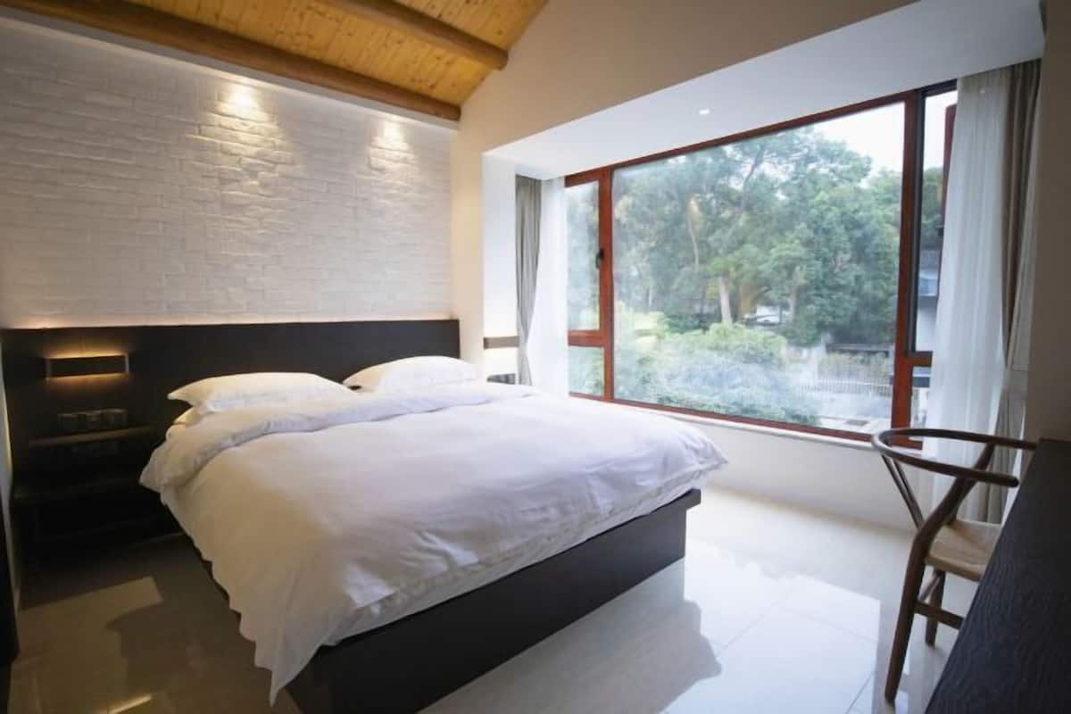 A modern bedroom with a large bed, a headboard with built-in lighting, a window with a view of trees, and a wooden ceiling.
