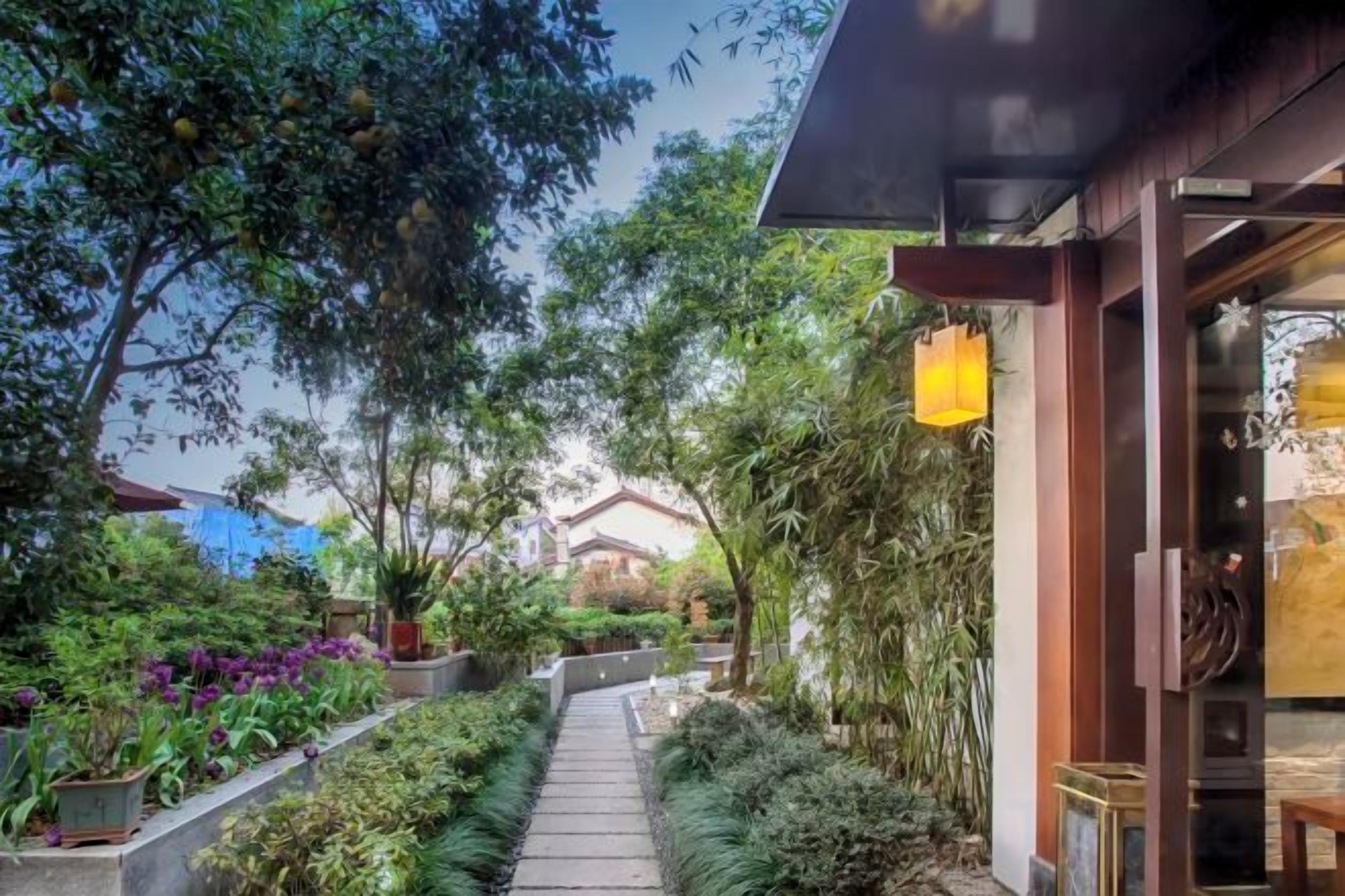 A pathway leading to a building with a covered entrance, surrounded by lush greenery and flowering plants.
