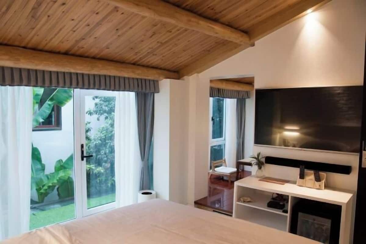 A bedroom with a bed, a TV, a desk, and a sliding glass door to a garden.
