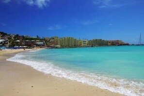 Ein Strand mit klarem türkisblauem Wasser, einem Sandstrand und einer Reihe mehrstöckiger Gebäude im Hintergrund.