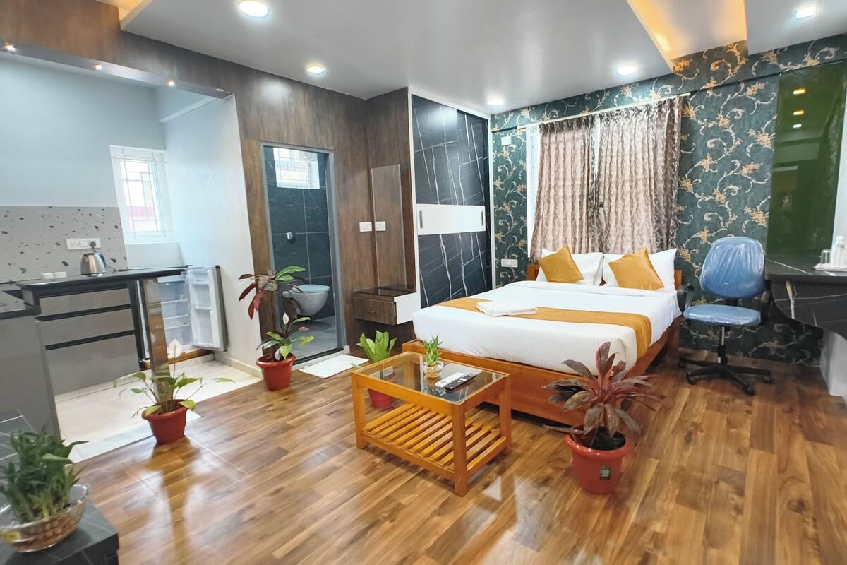 Une chambre d’hôtel moderne avec un plancher en bois, un lit avec une literie blanche et jaune, un bureau avec une chaise et plusieurs plantes en pot.