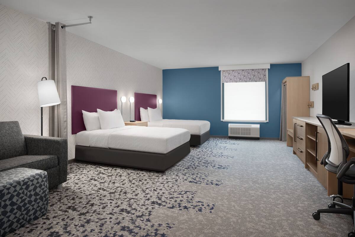 Ein Hotelzimmer mit einem Bett, zwei Nachttischen, einem Schreibtisch, einem Sessel, einer Lampe und einem Fernseher.