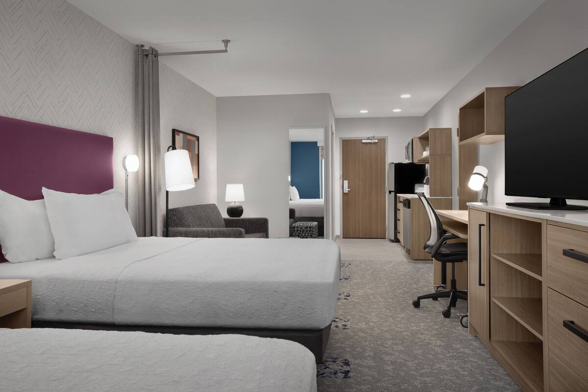 Ein Hotelzimmer mit zwei Betten, einem Schreibtisch, einem Fernseher und einem im Hintergrund sichtbaren Badezimmer.