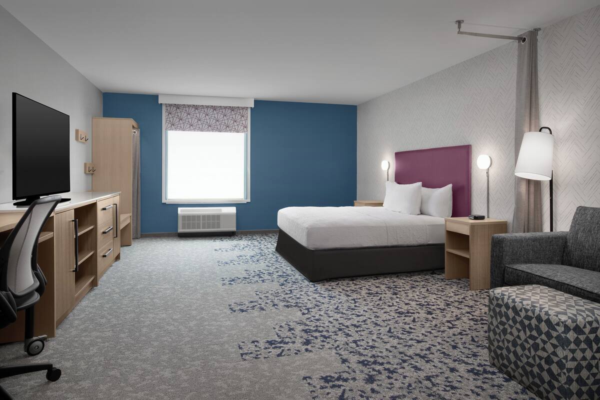 Ein Hotelzimmer mit einem Bett, einem Schreibtisch mit Fernseher, einem Sessel, einem Nachttisch mit Lampe und einem Fenster mit gemusterten Vorhängen.
