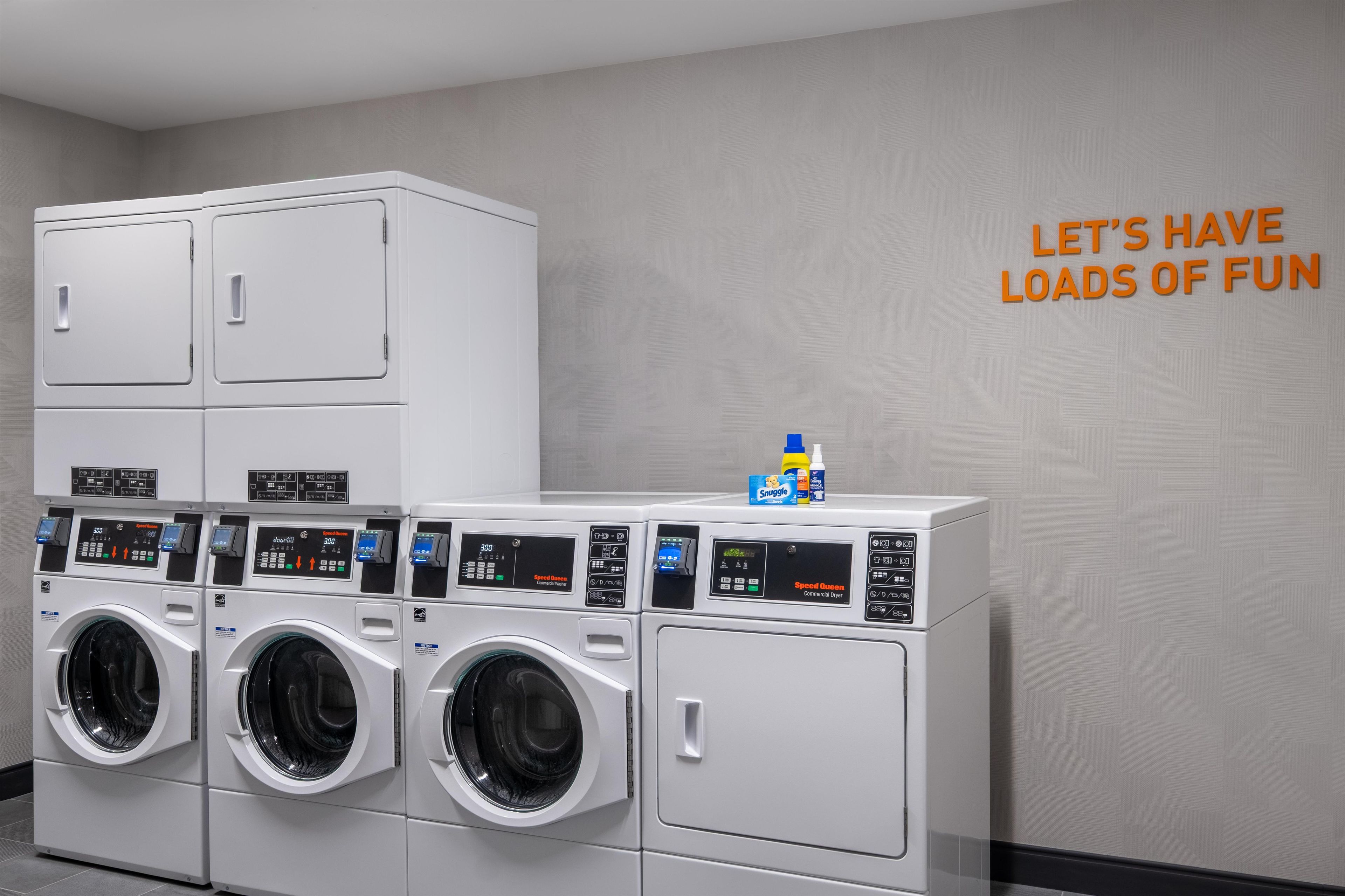 A laundry facility with multiple washing machines and dryers, a wall-mounted sign, and a bottle of detergent.