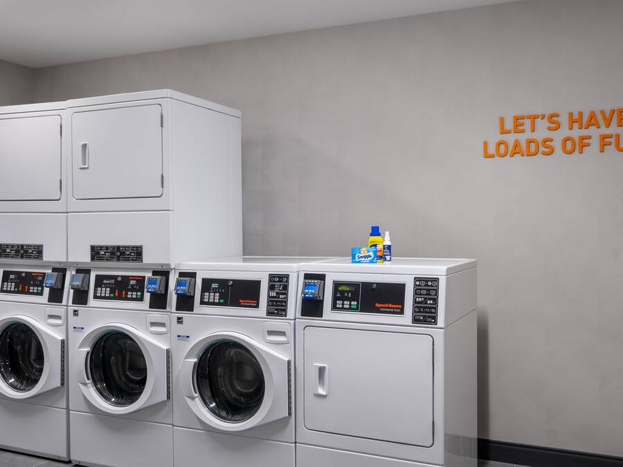 A laundry facility with multiple washing machines and dryers, a wall-mounted sign, and a bottle of detergent.