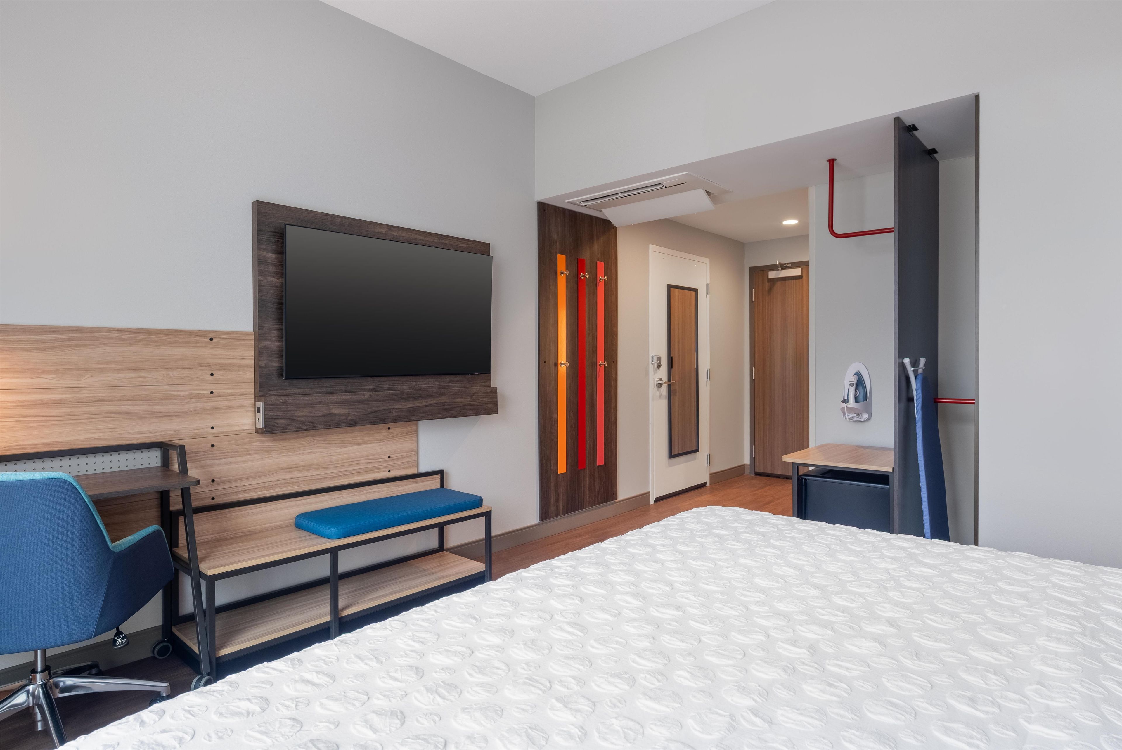 A hotel room with a bed, a desk with a chair, a television mounted on the wall, and a door with a red accent.
