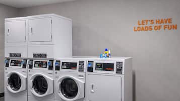 Una hilera de lavadoras y secadoras comerciales blancas en un cuarto de servicio con un letrero de pared que dice "LET'S HAVE LOADS OF FUN".