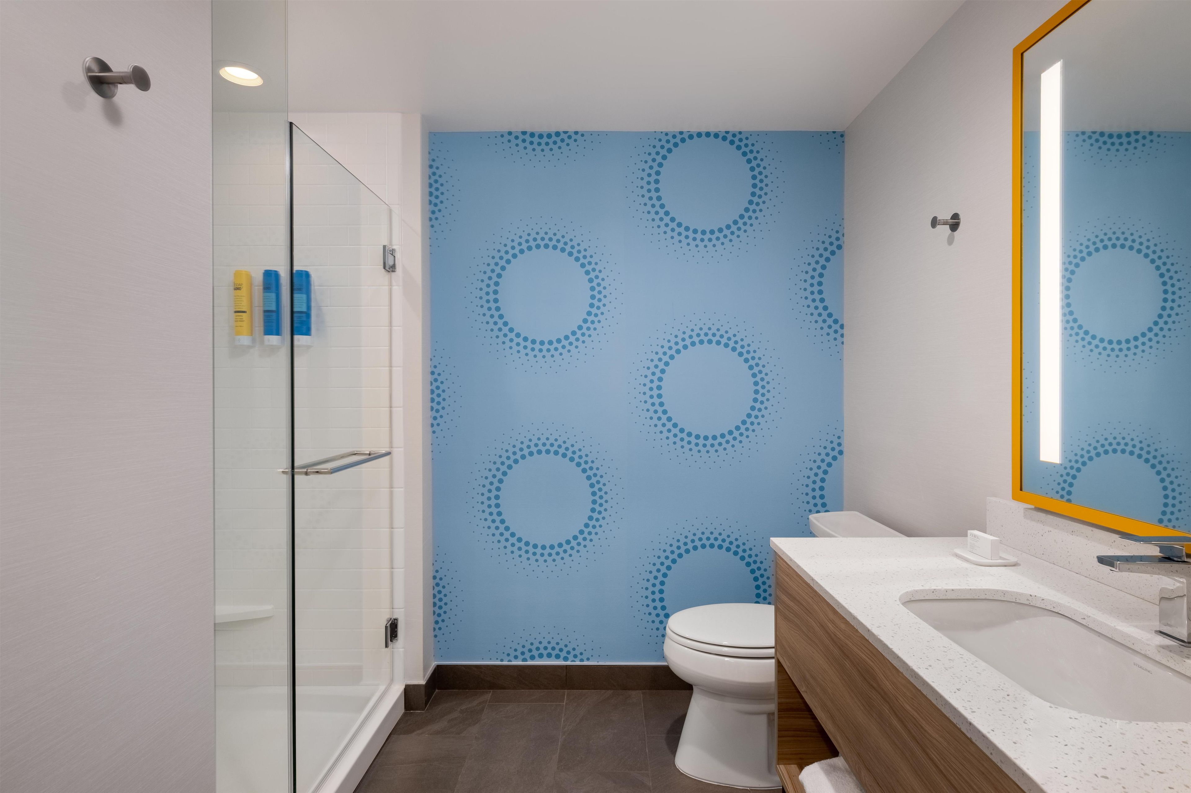 A bathroom with a glass shower enclosure, a toilet, a sink with a mirror, and a wall with a circular pattern.