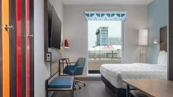 A hotel room with a bed, desk, chair, and a view of a building through a window.