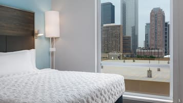 A hotel room with a large bed, a nightstand, a lamp, and a view of a cityscape through a large window.