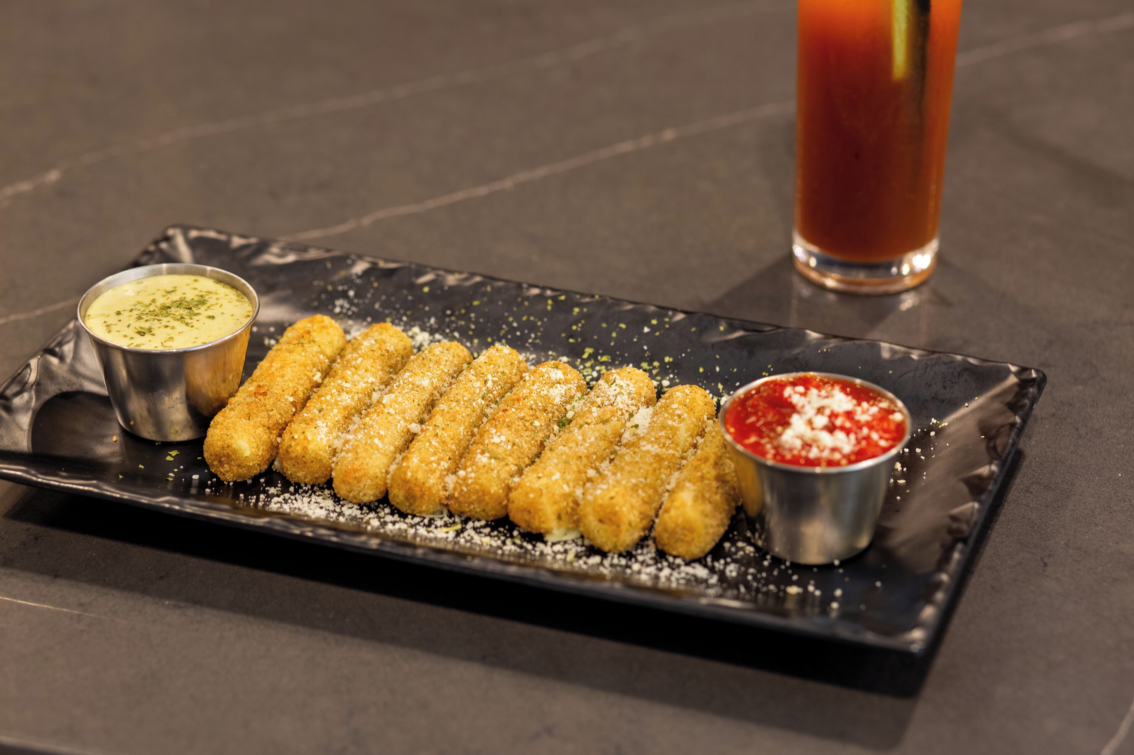 Une assiette rectangulaire noire contenant des aliments frits, deux sauces pour tremper et un grand verre d’une boisson rouge.