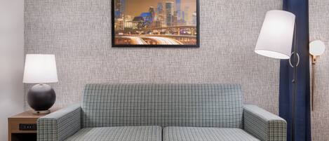 Una sala de estar moderna con un sofá estampado verde, un reposapiés rojo, un cuadro con una vista urbana enmarcada en la pared, una mesita de noche con una lámpara y una cortina azul.
