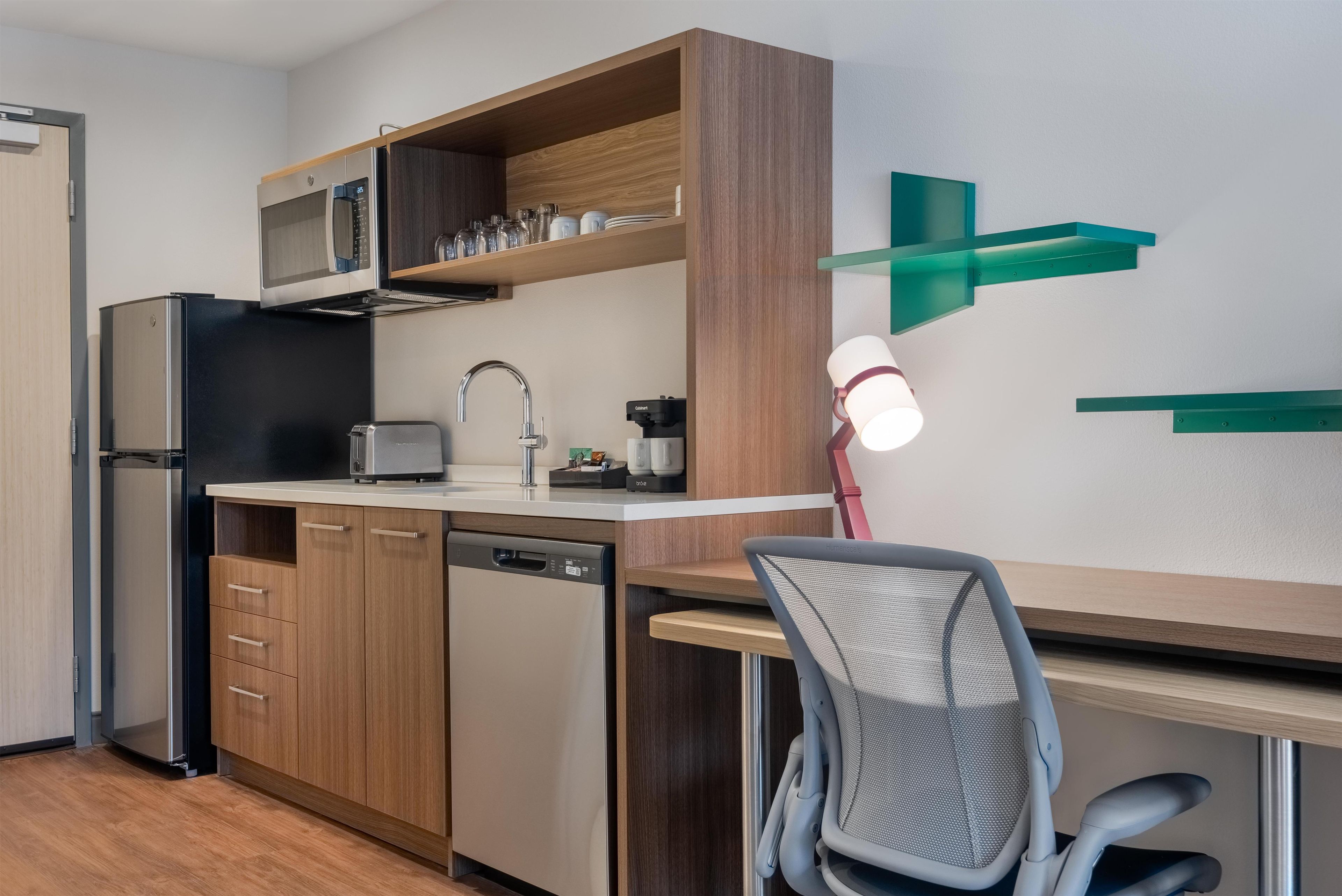 A compact kitchen with a refrigerator, microwave, and dishwasher, wooden cabinets, a small countertop, and a chair.