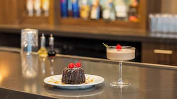 A dessert with cherries on a plate and a glass drink on a bar counter.