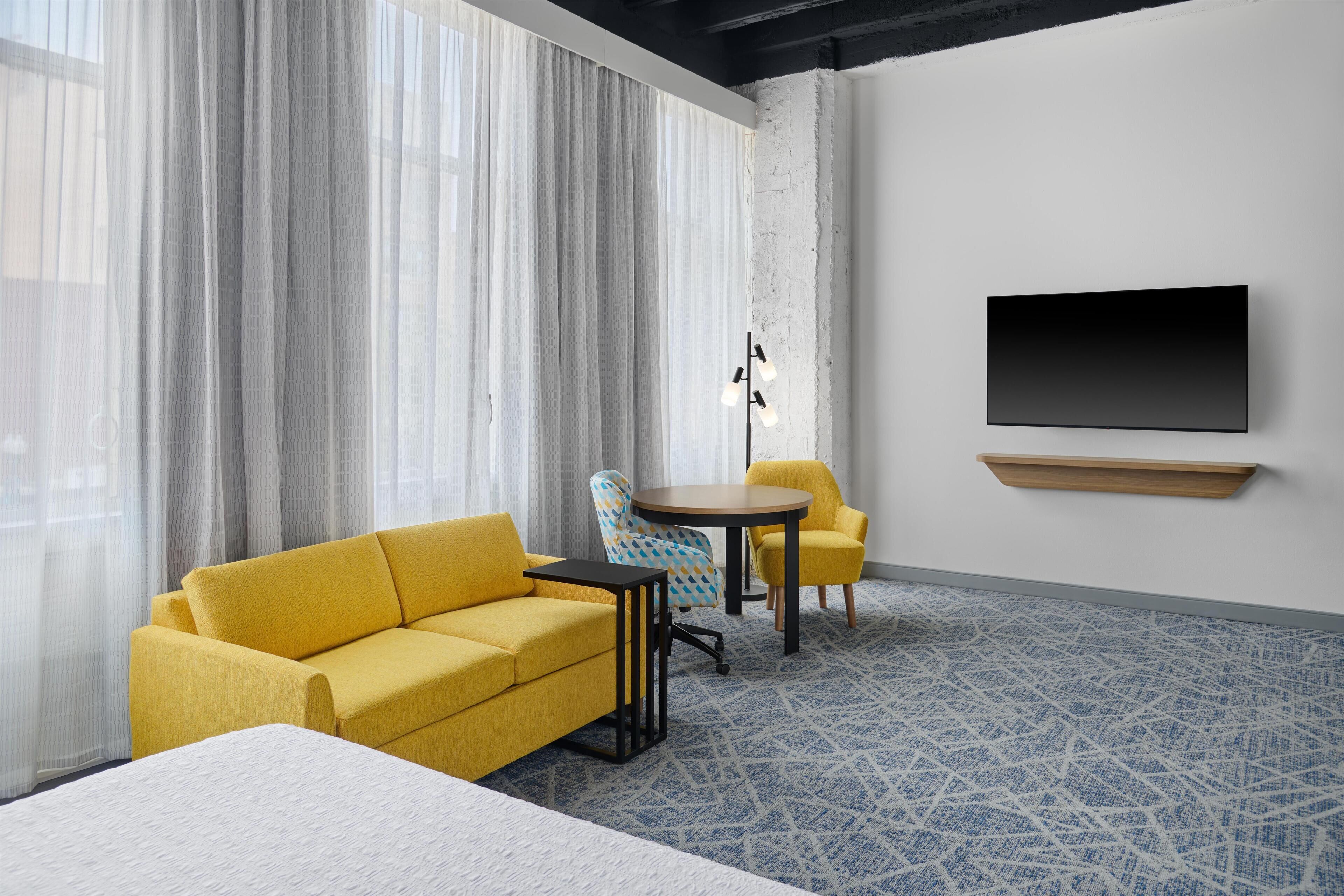 A hotel room with a yellow sofa, a round table, a TV mounted on the wall, and a patterned carpet.